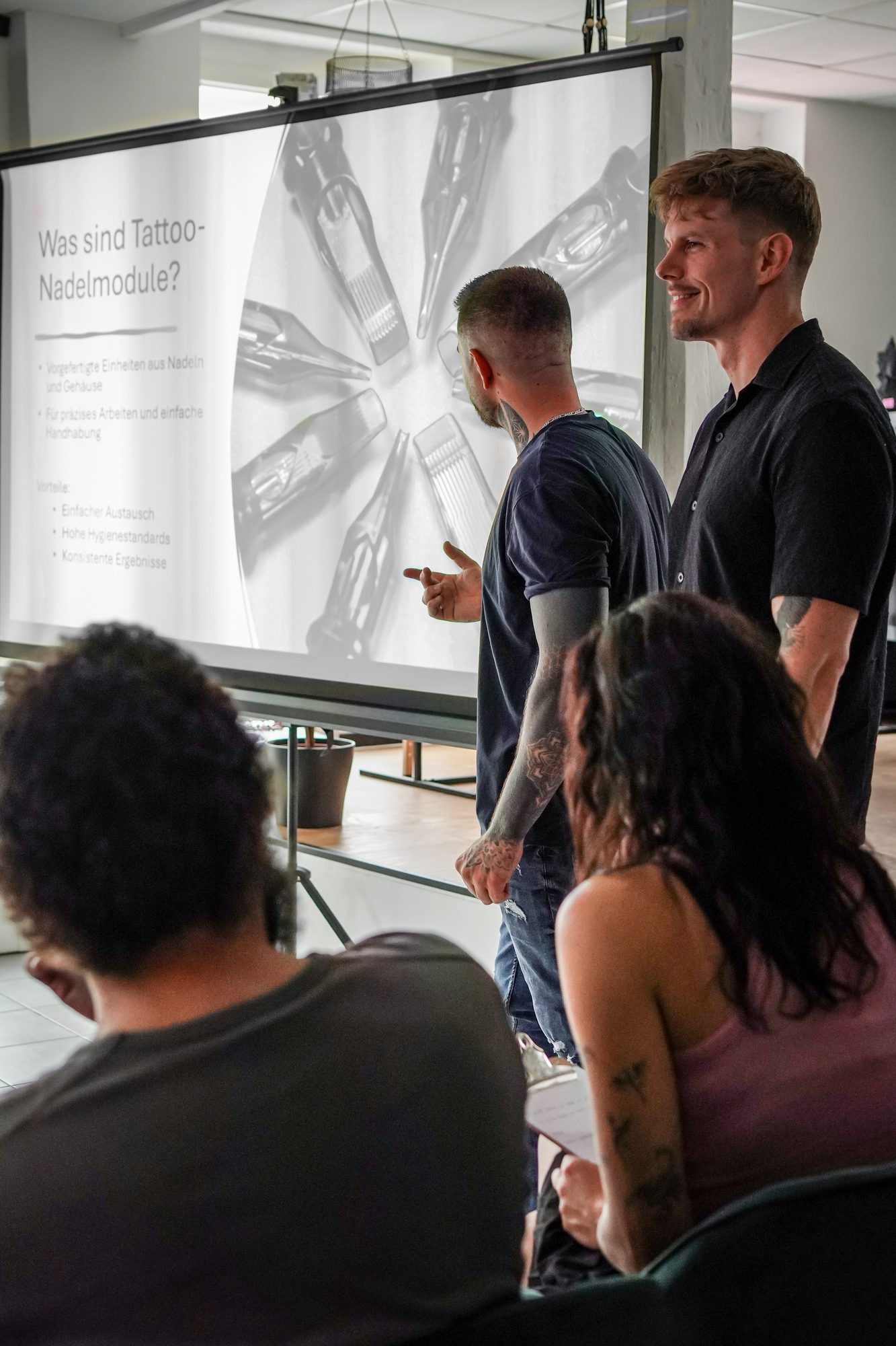 Zwei Trainer erklären Teilnehmern im Studio Backnang den Aufbau von Tattoo-Nadelmodulen anhand einer Präsentation